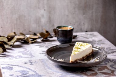 Çiğ vejetaryen peynirli kek, fırında glütensiz, limon aromalı ve fındıklı. Süslü seramik masada bir fincan sade kahve ve okaliptüs dalları. Güneş ışığı