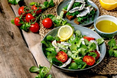 Yeşil tarla salatası turşulu ançüez ya da sardalya filetosu ve kiraz domatesli, limon ve zeytinyağlı mavi kasede servis edilen eski ahşap masa üzerinde saman peçetede..