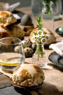 Renkli ve çikolatalı yumurtalar, sıcak haç çörekleri, buket çiçekleri, peçeteli boş seramik tabaklar, ahşap masa örtülü bir bardak limonata..
