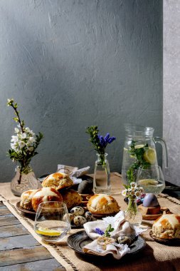 Renkli ve çikolatalı yumurtalar, sıcak haç çörekleri, buket çiçekleri, peçeteli boş seramik tabaklar, ahşap masa örtülü bir bardak limonata..