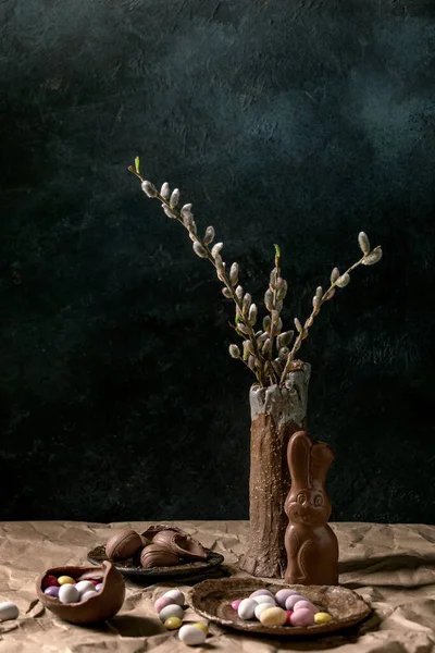 陶器の花瓶の花柳の枝 伝統的なチョコレートのウサギ 卵やお菓子とテーブルの上でイースター気分はまだ生活紙を砕いた 暗い背景 — ストック写真