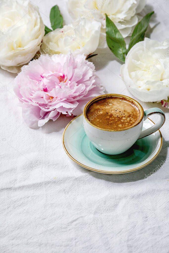 Cup of espresso coffee, pink and white peonies flowers with leaves over white cotton textile background. Flat lay, copy space