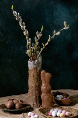 Paskalya havası seramik vazoda söğüt dalları, geleneksel çikolata tavşanı, yumurta ve şekerlerle dolu masada buruşuk el işi kağıtları. Koyu arkaplan.