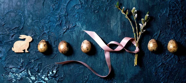 Çiçekli söğüt dalları, altın bıldırcın yumurtaları ve klasik mavi dokuya sahip tahta tavşanlı Paskalya tebrik kartı. Düz yat, boşluğu kopyala.