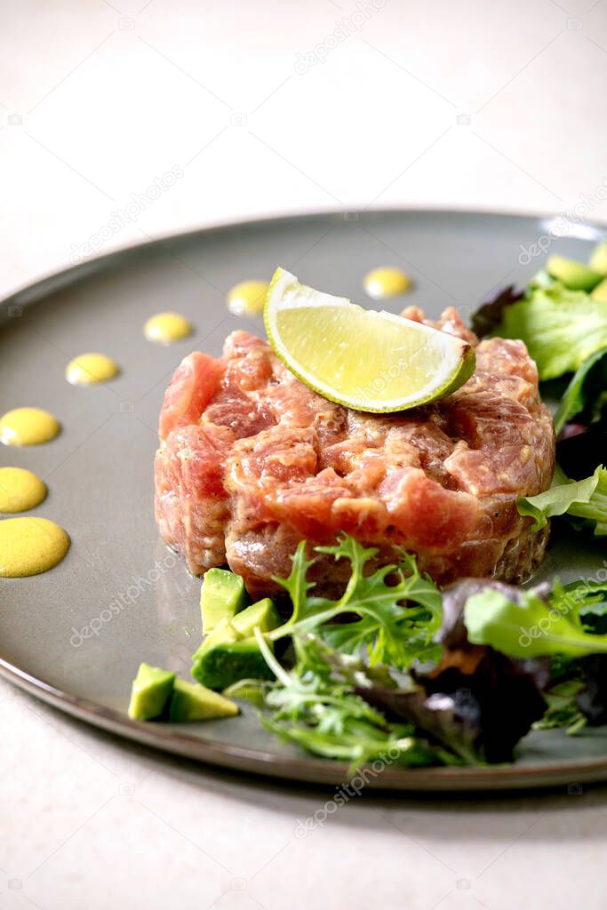 Tuna tartare with green salad, lime, avocado and mustard sauce serving on ceramic plate over white texture background. Fine dining, restaurant appetizer