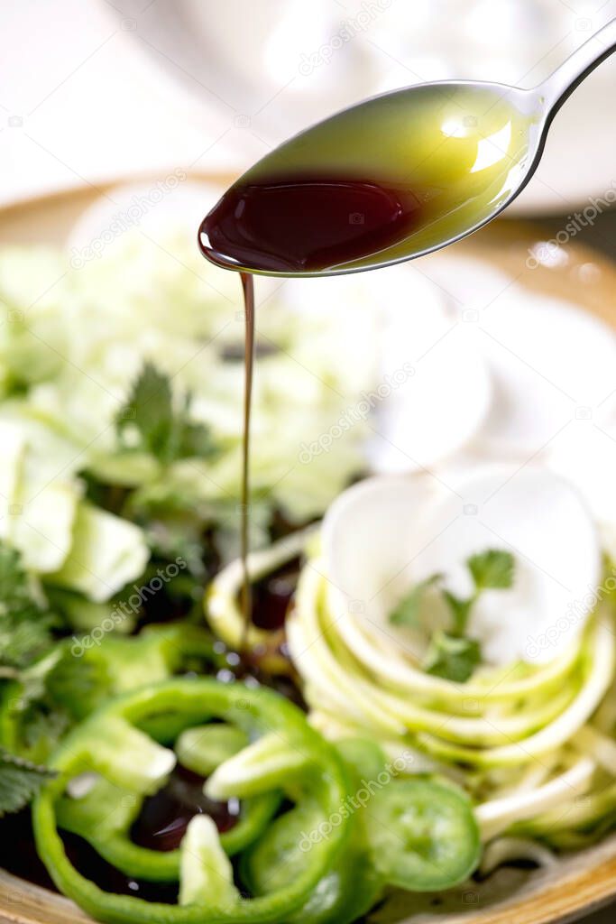 Pouring pumpkin seed oil on fresh green raw vegetables and herbs, spaghetti zucchini, white radish, green paprika, ice salad, mozzarella balls for cooking salad. Ceramic plate on white marble table.