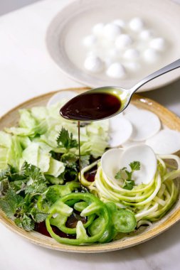 Taze yeşil çiğ sebze ve bitkilere kabak tohumu yağı, spagetti kabağı, beyaz turp, yeşil biber, buz salatası, pişirme salatası için mozzarella topları. Beyaz mermer masada seramik tabak.