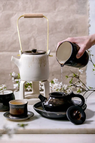 White and black handmade ceramic teapot with male hand pouring water from katakuchi jug, ceramic mugs, blossoming cherry tree as decoration standing on marble table. Traditional Japanese tea ceremony