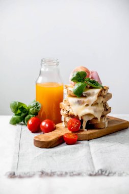 Kızarmış peynirli sandviç, jambon eti, kiraz domatesleri, portakal suyu ve fesleğen yaprakları beyaz mermer masa üzerinde ahşap kesme tahtasında.