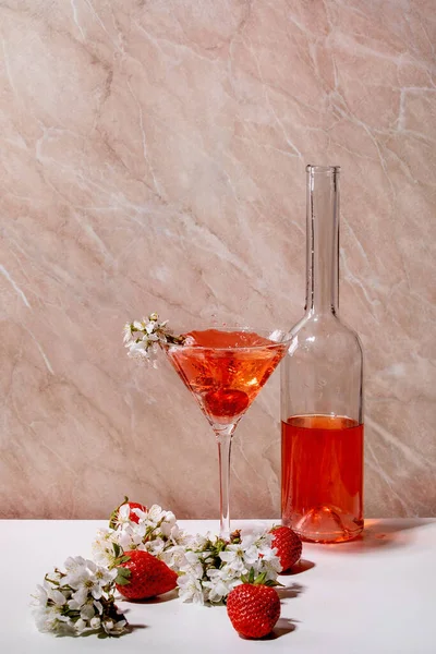 白とピンクの大理石を背景に桜の花の枝で飾られたマティーニグラスとボトルのストロベリーアルコールやノンアルコールカクテルの概念 — ストック写真