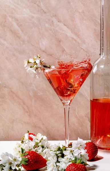 白とピンクの質感の背景に桜の花の枝で飾られたマティーニグラスとガラスボトルにイチゴアルコールやノンアルコールカクテルのスプラッシュ — ストック写真