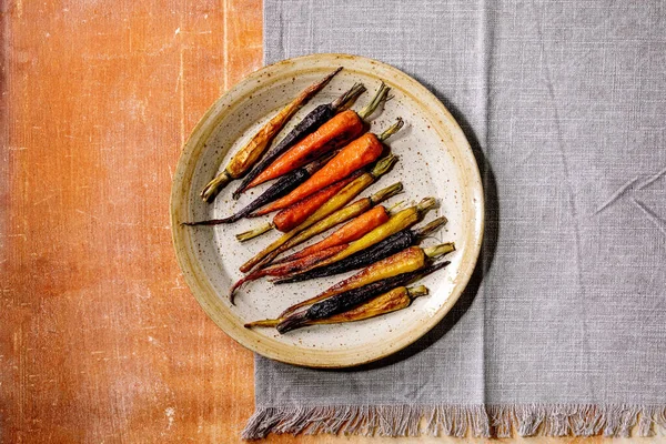 将不同颜色的胡萝卜烤在瓷盘上 用灰色的纺织品餐巾纸覆盖在橙色石头的背景上 复制空间 吃个饭 — 图库照片