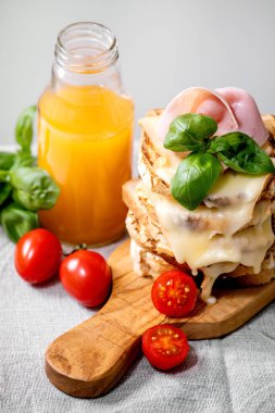 Kızarmış peynirli sandviç, jambon eti, kiraz domatesleri, portakal suyu ve fesleğen yaprakları beyaz mermer masa üzerinde ahşap kesme tahtasında.