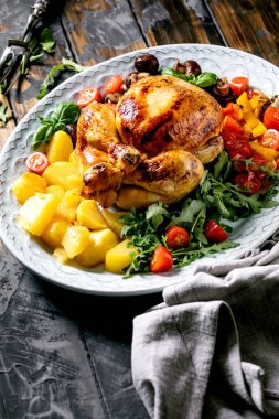 Izgara tavuk, sebzeli garnitürlü dolmalık biber, soğan, fırında patates, kiraz domatesleri, mantarlar ve siyah ahşap arka planda kumaş peçeteyle seramik tabakta otlar..