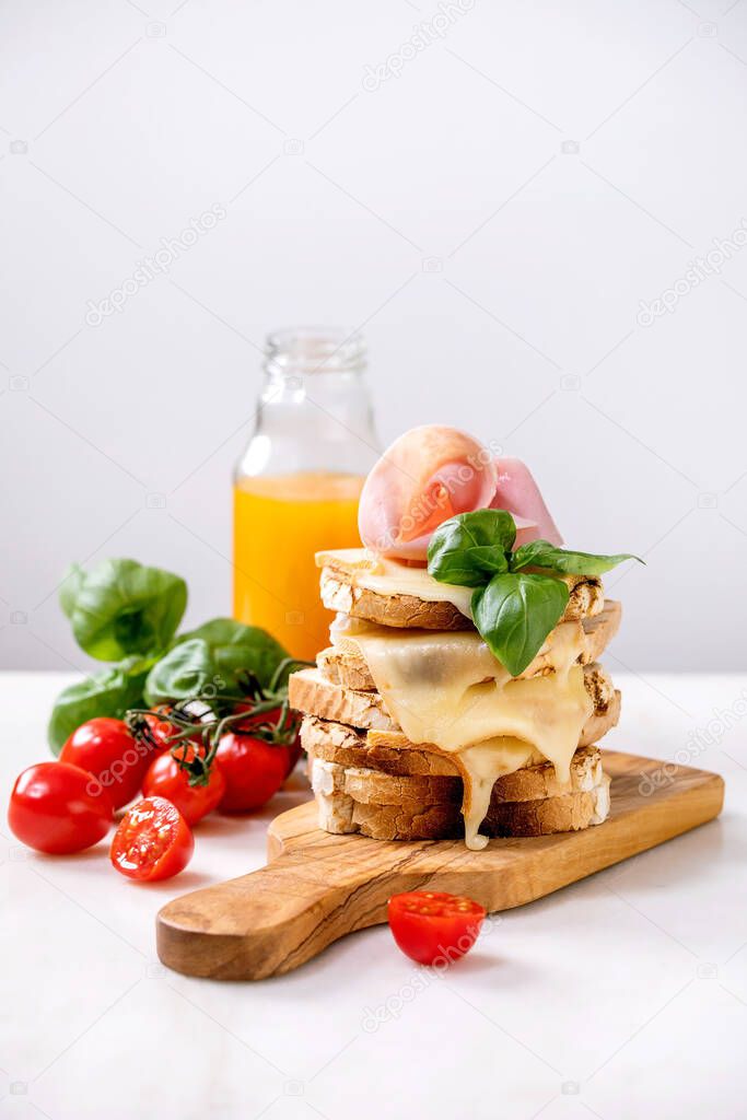 Stockpile of toasted melted cheese pressed sandwiches with ham meat, cherry tomatoes, orange juice and basil leaves on wooden cutting board on white marble table