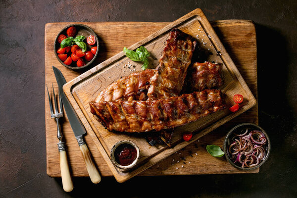 Grilled pork bbq ribs served with marinated onions, cherry tomatoes, basil and barbeque sauce on wooden cutting board over dark brown texture background. Vintage cutlery. Top view, flat lay