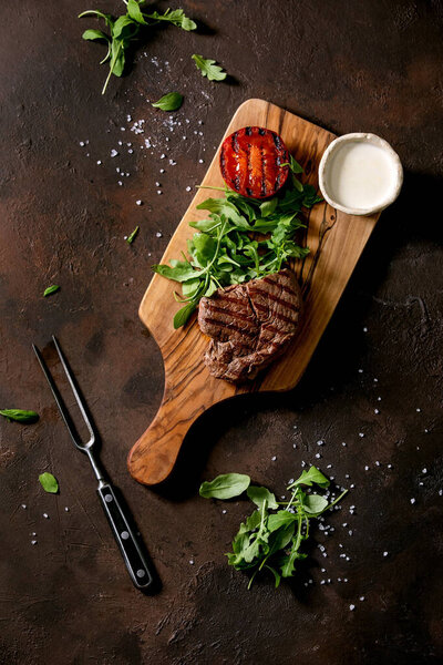 Grilled beef steak served with creamy sauce, grilled vegetables tomato and arugula salad on wooden cutting board with meat fork over dark brown texture background. Top view, flat lay