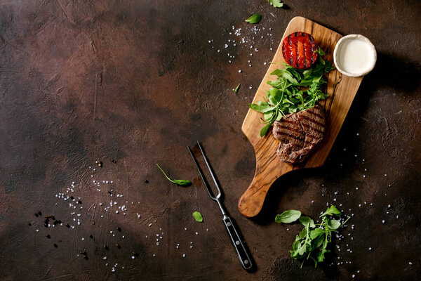 Grilled beef steak served with creamy sauce, grilled vegetables tomato and arugula salad on wooden cutting board over dark brown texture background. Top view, flat lay. Copy space