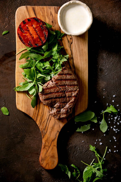 Grilled beef steak served with creamy sauce, grilled vegetables tomato and arugula salad on wooden cutting board over dark brown texture background. Top view, flat lay