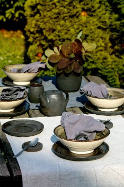 Bahçede boş seramik masa takımları, siyah tabaklar ve kaba kaseler ve eski ahşap masanın üzerinde keten masa örtüsü olan kırsal bir masa. Bahçe partisi