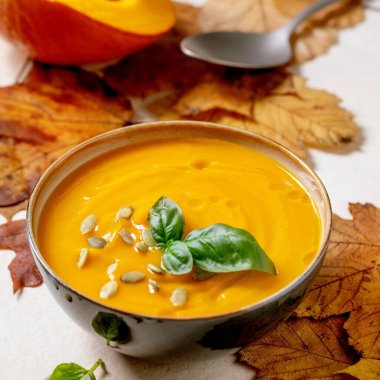 Bir kase balkabağı ya da taze fesleğen, zeytin yağı ve balkabağı tohumlarıyla süslenmiş havuç kremalı çorba. Beyaz dokuda sarı sonbahar akçaağaç yaprakları var. Kare resim