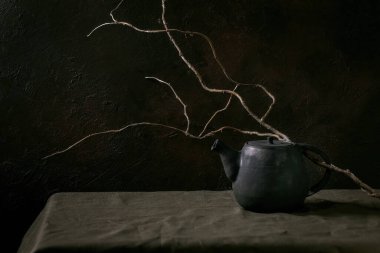 Çay seremonisi için el yapımı siyah seramik çaydanlık. Karanlık odada kuru dalla keten masa örtüsünün üzerinde duruyor. Boşluğu kopyala