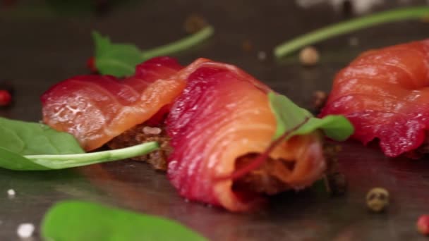 Sliced salmon filet, salted with beetroot juice, served on whole wheat toasts with salad leaves, sea salt and pepper over metal surface. Side view — Stock Video