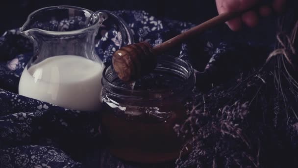Frasco de vidrio abierto de miel líquida con panal y jarra de miel en el interior, jarra de vidrio de leche y manojo de lavanda seca. Estilo rústico oscuro. En efecto filtro retro — Vídeos de Stock