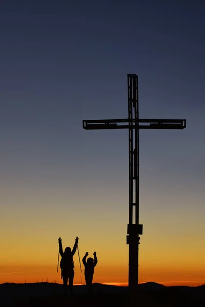 十字架の近くの山の上に子供を持つお母さんは勝利で腕を上げます — ストック写真