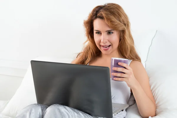 Jonge mooie vrouw met grappige gezicht zittend op haar slaapkamer met laptop en kopje thee — Stockfoto