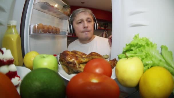音楽を聞いている男が、食べ物を求めて冷蔵庫を調べている。. — ストック動画