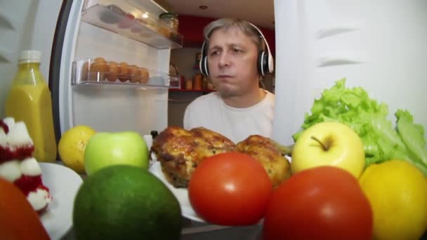 Seorang pria, mendengarkan musik, melihat ke dalam lemari es untuk mencari makanan. Sebuah kulkas sarjana penuh dengan makanan. — Stok Video