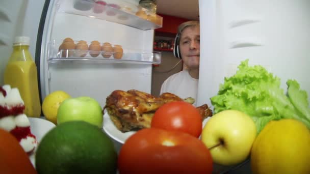 Um homem, ouvindo música, olha para a geladeira em busca de alimentos.Um refrigerador de solteiro cheio de comida. — Vídeo de Stock