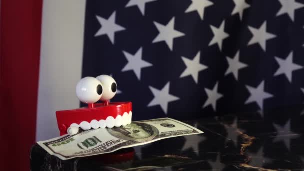 Uma prótese de brinquedo segura uma nota de dólar em seus dentes contra o fundo da bandeira americana . — Vídeo de Stock