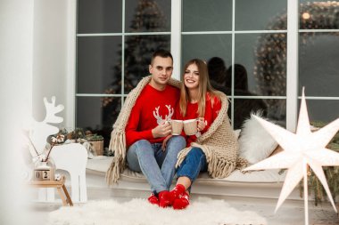 Noel 'i birlikte kutluyoruz. Panoramik pencerelerin yanındaki pencere eşiğinde genç ve güzel bir çift Noel arkaplanından kameraya gülümsüyor..