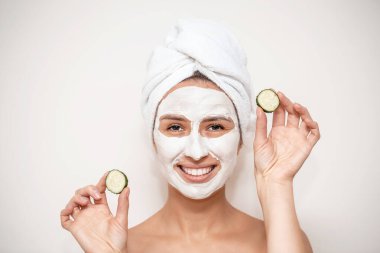 Kafasına havlu sarılmış, maske takmış, elinde salatalık dilimleri olan güzel genç bir kadın. Yüz maskesi, cilt bakımı ve tedavi konsepti.