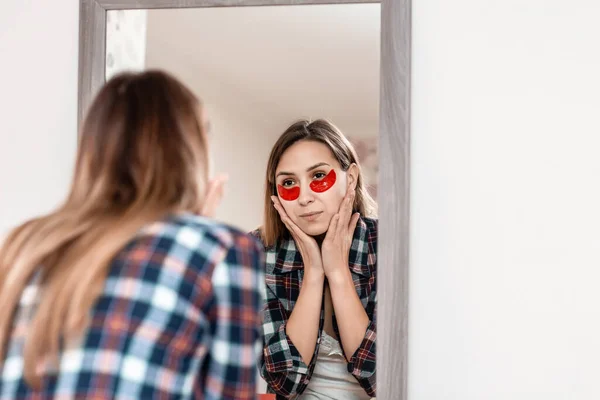 Beautiful woman applying anti-fatigue under-eye mask watching in the mirror. Skin care girl touch patches under eyes to reduce eye bags. Spa procedures at home.