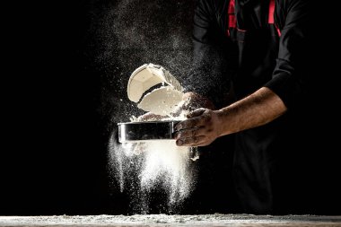 Şef eleğe un serpiyor, toz un havaya karışıyor. Un bulutunun dondurucu hareketinde un taşıyan şefin elleri.