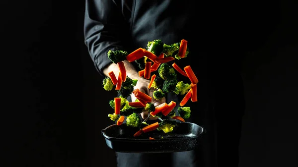 flying vegetables scattering in a freeze motion of a cloud midair on black. Cook hands with vegetables on a pan. banner, catering menu recipe place for text.