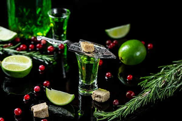 absinthe in glass with lime slices, sugar cubes, cranberries and lime, stainless steel spoon on dark background.