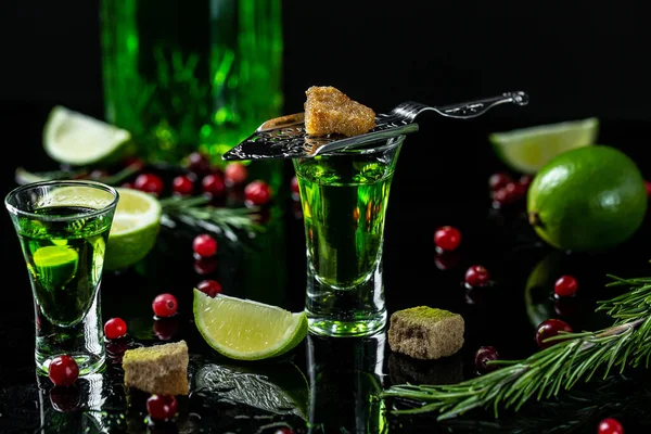 a glass of absinthe, green cocktail. stainless steel slotted spoon with the sugar cube cranberries and lime, the absinthe bottle.