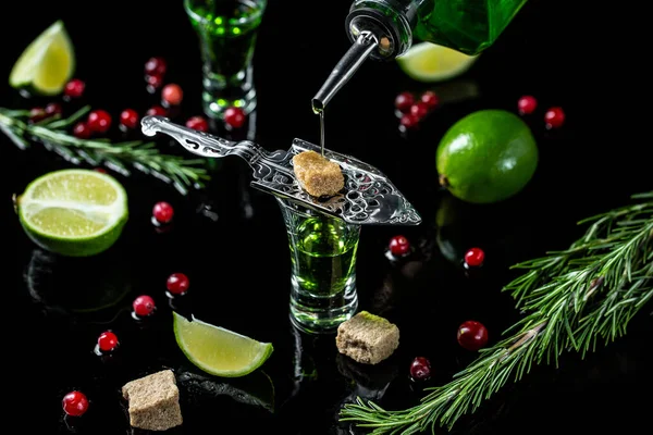 absinthe shots with sugar cubes. absinthe poured into a glass. bottle of absinthe with brown sugar, cranberries and lime, stainless steel spoon isolated on black background