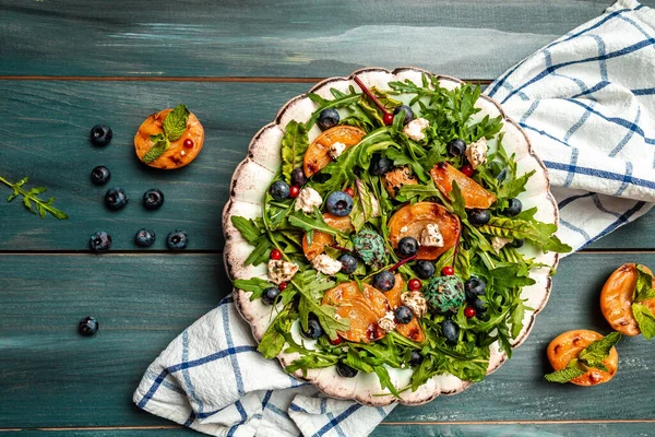 Easy recipe for summer salad with goat cheese, grilled apricots, arugula, berries, close-up on a plate wooden background, recipe top view.