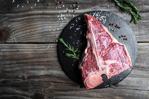 Raw T-bone Steak or porterhouse steak for grill or BBQ with spices and herbs on slate board and woden rustic background, top view, border.