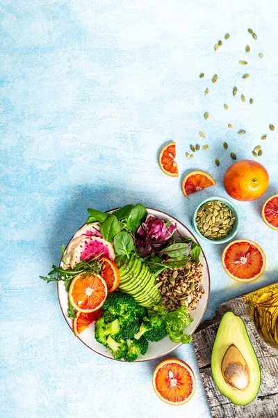 Veggie bowl salad with vegetables, avocado, blood orange, broccoli, watermelon radish, spinach, quinoa, pumpkin seeds on a blue background. vertical image, place for text