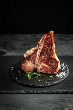 Çiğ kemik bifteği ya da siyah arka planda biftek, tuz, biber ve otların yanında. dikey resim, metin için yer.