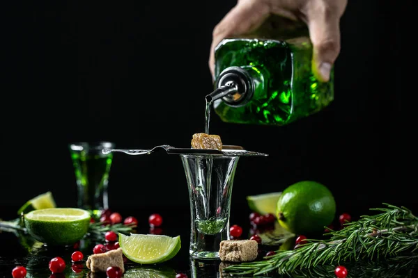 absinthe shots with sugar cubes. absinthe poured into a glass. bottle of absinthe with brown sugar, cranberries and lime, stainless steel spoon isolated on black background