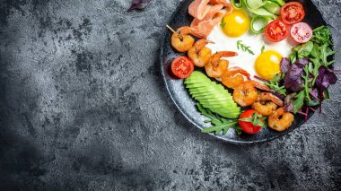 Keto kahvaltısı düşük karbonhidratlı somon, ızgara karides, karides, kızarmış yumurta, taze salata, domates, salatalık ve avokado. Uzun pankart biçimi, üst görünüm.