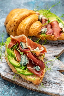 Jamon jambonlu kruvasan sandviçi serrano paleta iberica, mavi peynir, avokado, mavi arka planda mikrocin. Kahvaltı konsepti, yemek tarifi geçmişi. dikey resim.