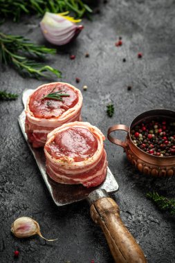 Çiğ mermer et, biftek fileminyonu. Koyu beton arka planda eski kasapta servis edilen domuz pastırmasına sarılı madalyon biftekleri. dikey resim, metin için yer.